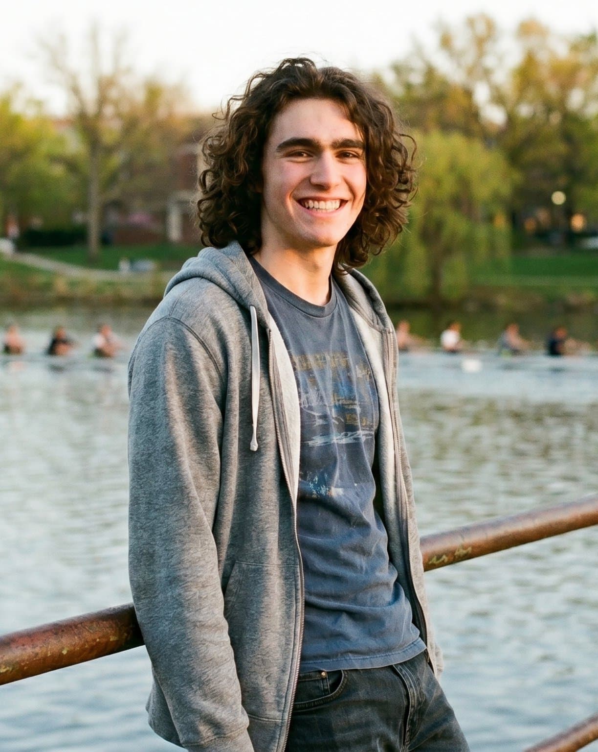 Ethan Highfield in a gray zip-up hoodie over a blue tee, leaning on a railing by the water in soft evening light, smiling.
